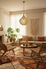 A large, organic-shaped pendant light made of woven natural fiber, casting a bright glare from the exposed bulb inside, hanging against a rustic brick wall with a framed art print behind it.